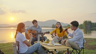 小组四人朋友亚洲男女野营，喝啤酒，庆祝，唱歌，弹吉他，玩乐，享受地面帐篷。日落假期期间的水库区域。视频素材模板下载