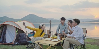 小组四人朋友亚洲男人和女人野营野餐庆祝，通过智能手机自拍享受乐趣和地面帐篷。日落假期期间的水库区域。