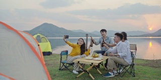 四人组朋友亚洲男女野营，喝啤酒，庆祝，通过智能手机自拍，享受地面帐篷。日落假期期间的水库区域。