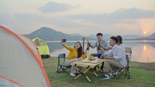 四人组朋友亚洲男女野营，喝啤酒，庆祝，通过智能手机自拍，享受地面帐篷。日落假期期间的水库区域。视频素材模板下载