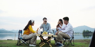 群亚洲四青年朋友男女在河边野餐野营，他们有说有笑，喝着庆祝欢呼啤酒。