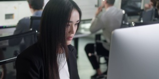 有吸引力的亚洲妇女在台式电脑上工作，而在大的开放空间办公室工作。正面商业女性的肖像看着室内的电脑屏幕