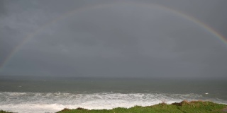 在彩虹下。在海上的海岸上，一道彩虹几乎完全可以看到在悬崖脚下，海浪拍打着海岸，形成白色的泡沫，海鸥在它周围飞翔