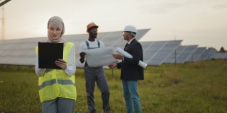 戴头巾的妇女在太阳能电站工作。戴着头巾的妇女站在太阳能电池板上看着相机。技术员和印度工程师进行背景讨论