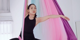 关注健康的年轻亚洲妇女在黑色运动服做瑜伽飞行与晾衣绳姿势空中使用彩色吊床。空中反重力瑜伽女子用彩带表演瑜伽飞行。柔韧性，集中力，反重力平衡。