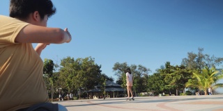 在阳光明媚的日子里，一个亚洲男孩试图给他的妹妹在公园玩滑板的时候拍照。