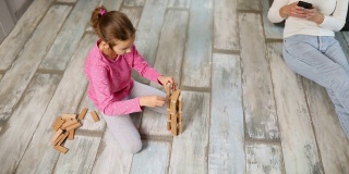 无所谓的母亲用手机，女儿在家里的地板上玩木结构