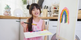 亚洲年轻快乐的小女孩在客厅的画板上画画。可爱的小朋友在家里学习如何用水彩画和毛笔画画，享受假期的创意活动。