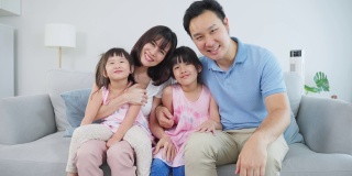 亚洲幸福家庭的肖像，花时间与孩子一起在家里。年轻可爱的小女孩和父母坐在沙发上微笑，看着客厅里的镜头。活动关系的概念