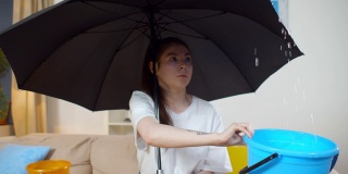 一名困惑的女子坐在客厅的伞下，拿着水桶准备接天花板漏水的水