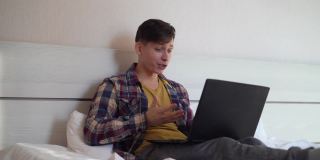 年轻的白人男子坐在家里的床上，穿着便服，用他的笔记本电脑视频聊天。C