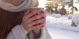 在阳光明媚的寒冷的冬天，在阳光的背景下，不认识的年轻女子的特写镜头握着杯子热气腾腾的饮料。