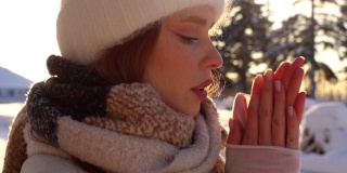 特写脸冻僵的年轻女子在冬天外套等待在寒冷的风天气，搓手，吹手掌保暖。