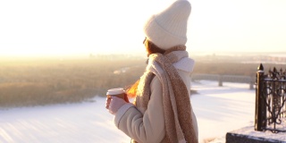 幸福微笑的年轻女子站在观景台上看日落，手里拿着一杯热咖啡。