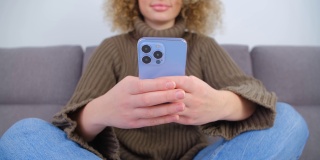 在封锁期间，一名年轻女子坐在家中客厅的沙发上，用带有三个摄像头的现代蓝色手机打字。4k视频中的互联网传播娱乐理念
