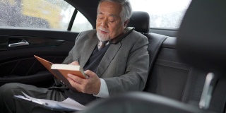 4K亚洲资深商人坐在汽车后座，一边打电话一边在书中写商业计划