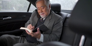 4K亚洲资深商人坐在汽车后座，一边打电话一边在书中写商业计划