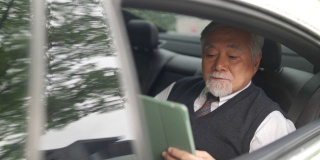 4K亚洲资深商人坐在汽车后座，一边喝着热咖啡，一边在数码平板电脑上工作