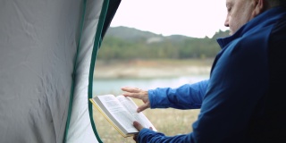 老人在帐篷里看书，在山前湖边露营——老人在大自然的概念下旅行