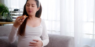 亚洲妇女怀孕期间在家里的沙发上吃不健康的披萨。垃圾食品