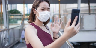 慢动作场景:年轻的亚裔员工女性，早上戴着口罩站在公共交通入口处，用智能手机上班，追求新常态和健康的生活方式理念