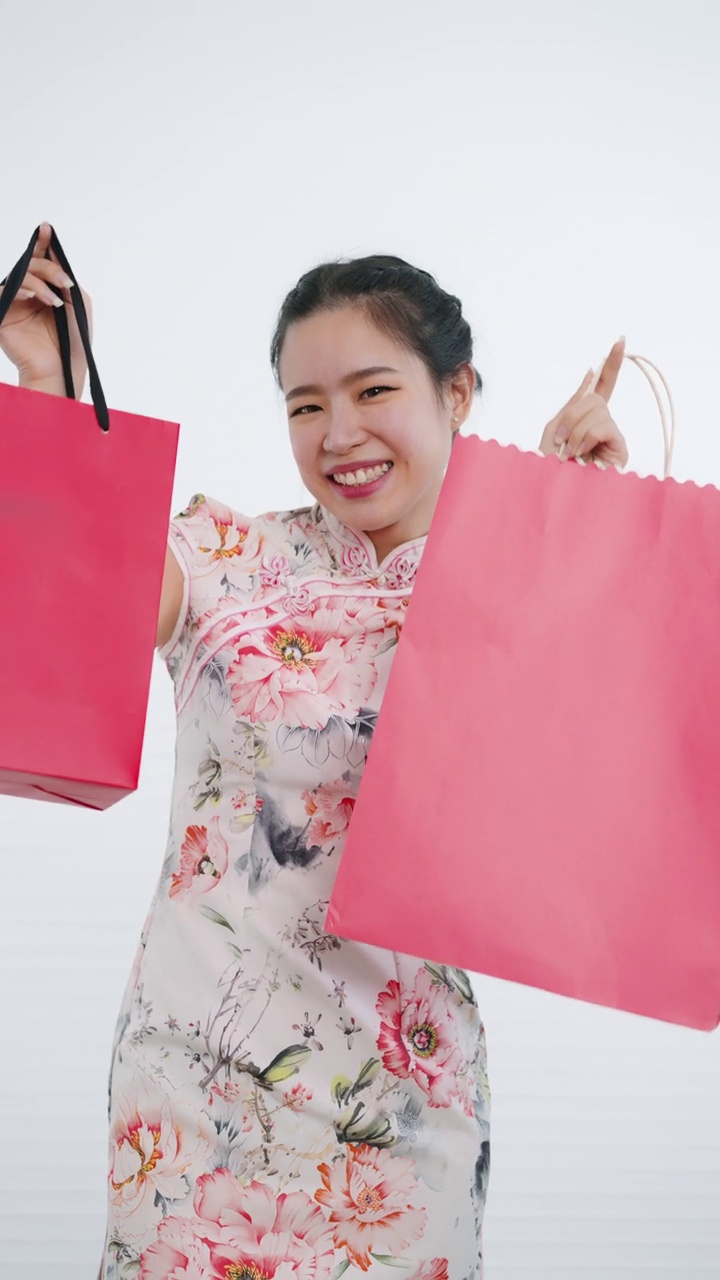 中国/亚洲妇女在中国新年期间，双手拿着红色和粉红色的购物袋，白色背景，穿着旗袍白色花卉图案连衣裙