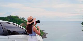 年轻的女性旅行者看着和拍照在美丽的海景与她的车，而旅行驾驶公路旅行的假期