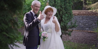 快乐兴奋的高级新娘和新郎的肖像手势大拇指向上微笑着看着相机。快乐的白种人男人和女人在婚礼当天在户外摆慢动作。幸福和关系。