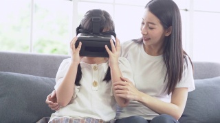 亚洲母女坐在客厅的沙发上，戴着虚拟现实(vr)眼镜，面带微笑地玩着电子游戏。幸福家庭元虚拟现实技术概念。视频素材模板下载