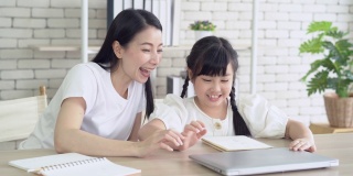 做得很好。年轻的亚裔母亲兴奋地击掌庆祝女儿完成了家庭作业。快乐的妈妈和小女孩在家快乐的学习。教育，在线学习的概念。