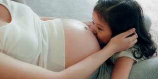 怀孕的母亲和学龄前的女儿坐在家里的沙发上。可爱的女孩亲吻怀孕妈妈的肚子。