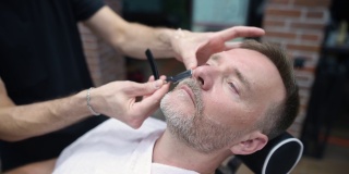 理发师大师在沙龙用电动剃须刀为英俊成熟的大胡子男子剃须。发型艺术家为男士理发店的人制作胡须造型。专业造型师服务。