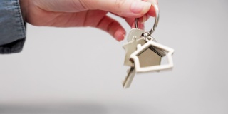 一个女人穿着衬衫的手抖动着金属钥匙链房子的新钥匙的特写镜头。灰色的背景。抵押、租赁和房地产的概念