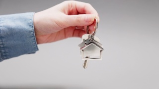 一个穿着衬衫的女人的手的特写清晰地显示了一个金属钥匙链房子的新钥匙。灰色的背景。抵押的概念，购买房地产和租赁视频素材模板下载