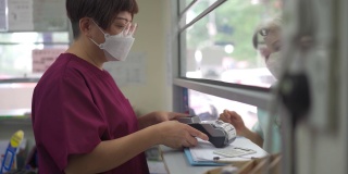 亚裔华裔老年妇女戴着口罩，在诊所用信用卡刷卡，女护士拿着信用卡读卡器