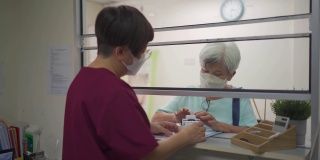 亚裔华裔老年妇女戴着口罩，在诊所用信用卡刷卡，女护士拿着信用卡读卡器