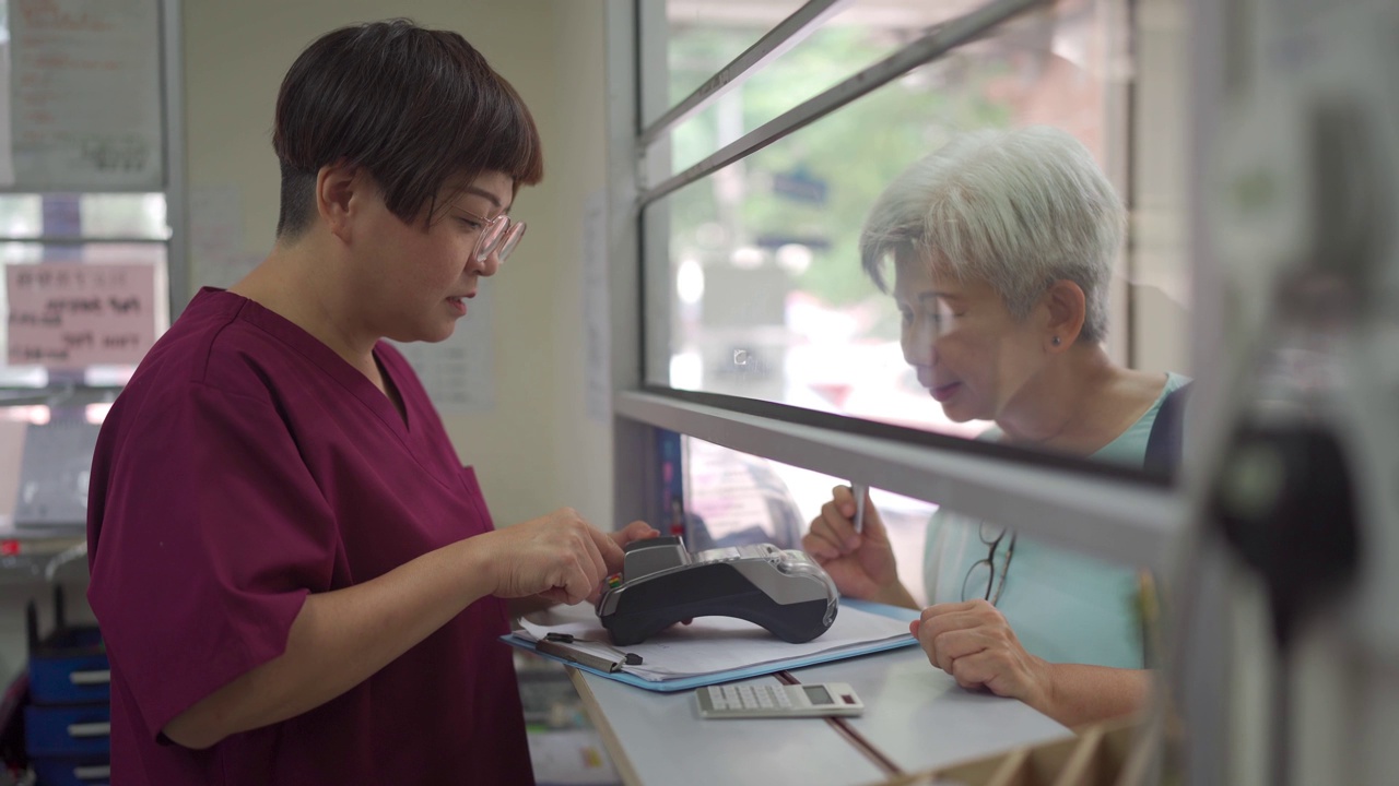 亚洲华人老年妇女用信用卡支付医药费，女护士手持信用卡读卡器在诊所刷卡