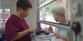 亚洲华人老年妇女用信用卡支付医药费，女护士手持信用卡读卡器在诊所刷卡