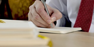 一个用钢笔在笔记本上书写的手的特写。男人坐在桌子上看书。