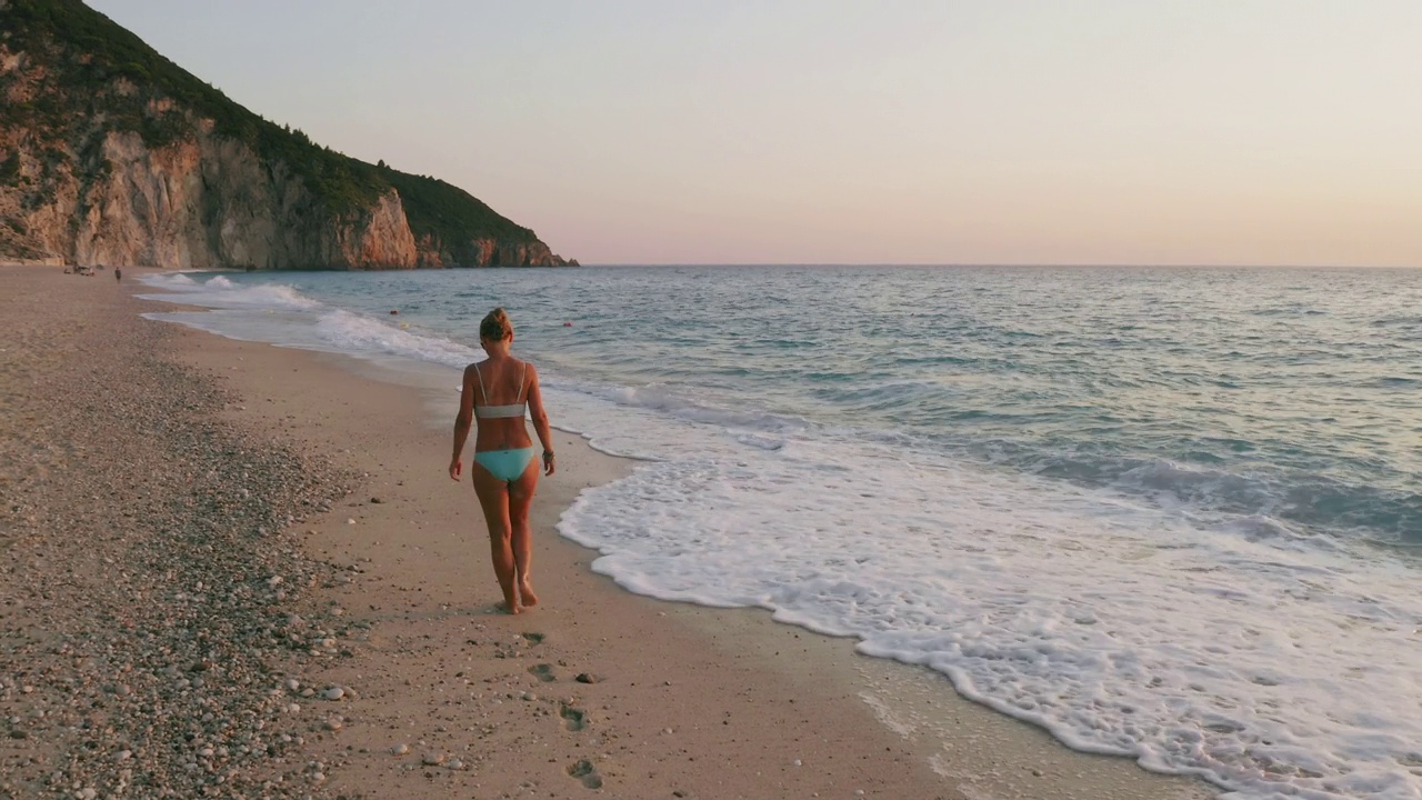 空中低空跟随成年妇女享受美丽的米洛斯海滩的Lefkada，爱奥尼亚岛，希腊。夕阳金色的阳光和原始的海浪打破了蓝绿色的大海在岸边