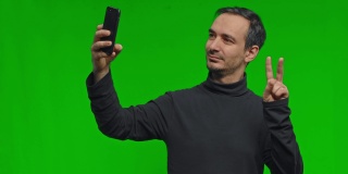 一名男子正在用手机自拍，在孤立的绿色背景上摆出胜利的姿势