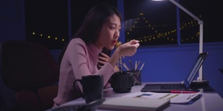 亚洲商界女性晚上在家边吃方便面边用笔记本电脑工作