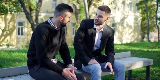 两个自信的留着胡子的年轻白人男子坐在城市公园的户外长椅上聊天。阳光明媚的周末，帅气的男性朋友在闲聊。休闲的生活方式。