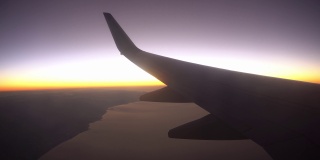 飞机在日落或日出时飞越大海和沙漠，从飞机的机翼和襟翼的窗口看到的景色。