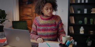 在线教育年轻卷曲的非裔美国妇女做项目研究，做笔记，喝咖啡在家里