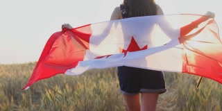 年轻快乐的加拿大女孩在麦田里张开双臂奔跑，在日落时举着加拿大国旗。慢动作