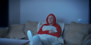 一个穿着连帽运动衫的男人，晚上坐在沙发上，把脚放在前面看电视，电视发出的光