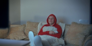 穿着红色连帽运动衫的男人，晚上坐在沙发上把脚放在面前看电视，电视灯