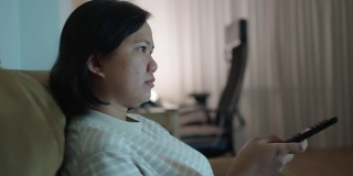 亚洲妇女坐在沙发上选择电视频道的遥控器在晚上看