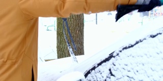 一名男子在雪后从车上刷雪。一只穿着芥末色夹克的手，白色的身体上放着汽车扫把。冬天的天气条件。慢动作